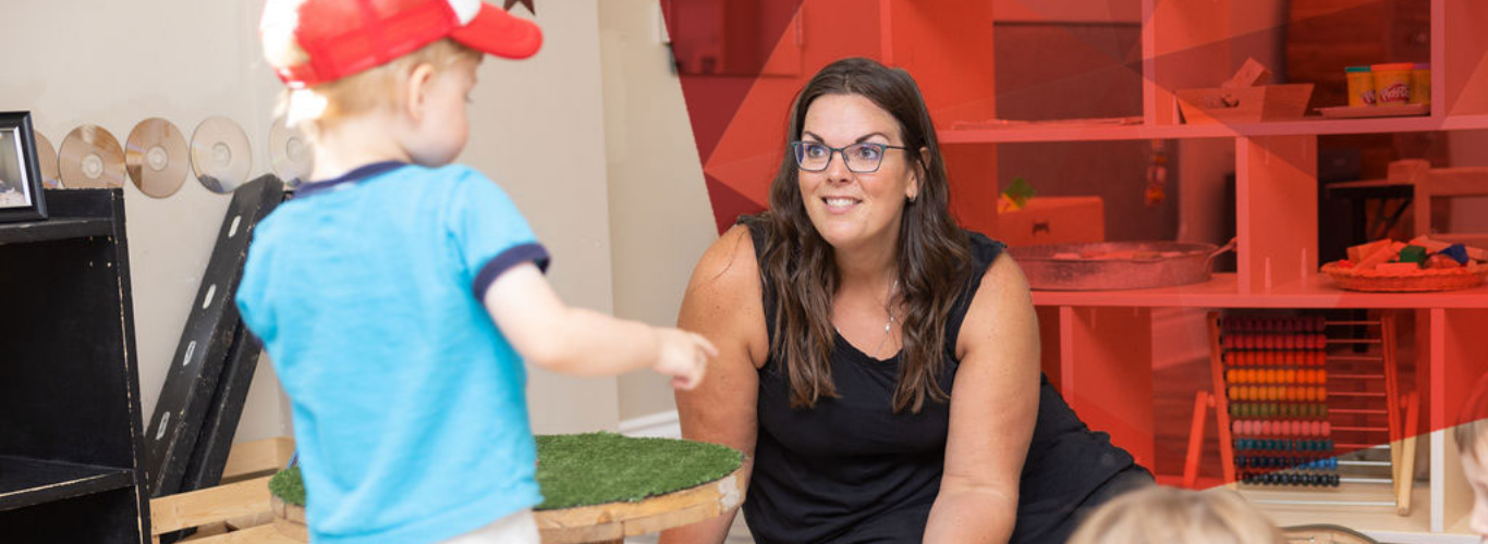 A woman in a daycare setting smiles at a toddler in a red hat